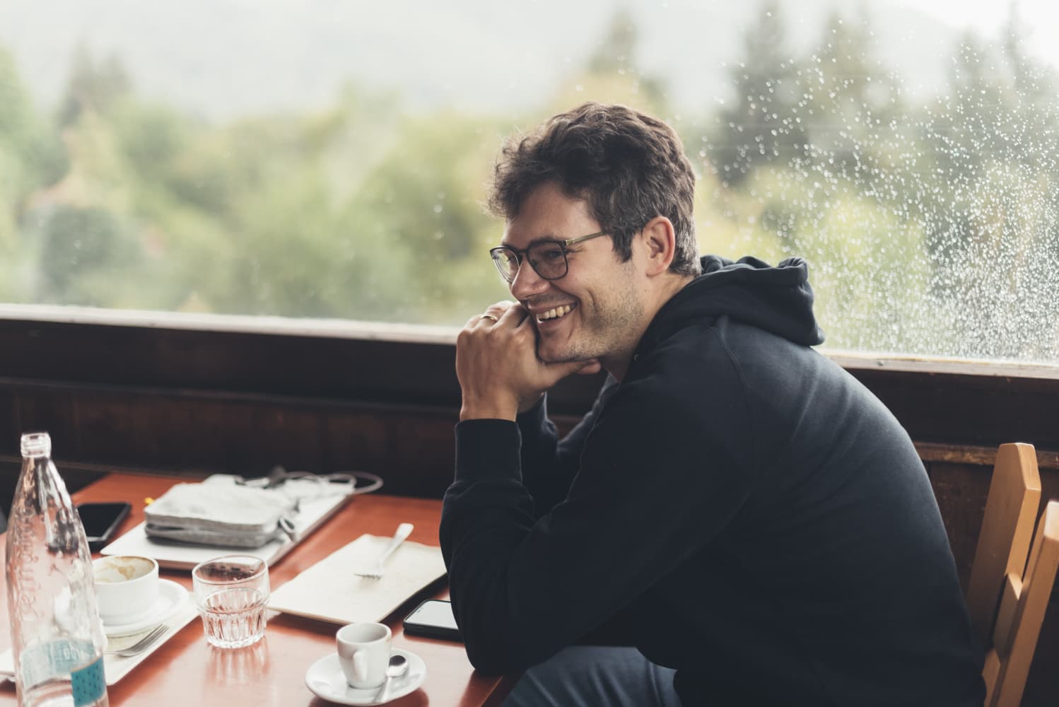 Un uomo sorridente seduto a un tavolo accanto a una finestra coperta di pioggia.